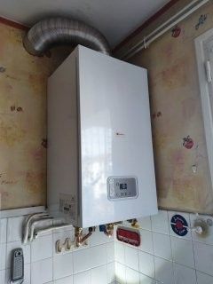 Chaudière à gaz murale blanche avec affichage numérique, tuyau d'évacuation et plomberie contre un mur carrelé.