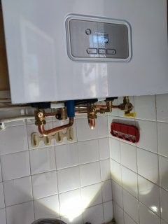 Chaudière à gaz blanche fixée sur un mur carrelé, avec tuyaux en cuivre et panneau de commande.