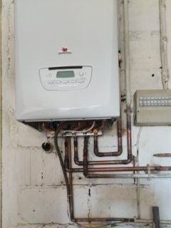 Chaudière à gaz murale blanche avec tuyaux en cuivre, affichage numérique et petit tableau électrique.