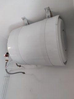 Chauffe-eau blanc fixé sur un mur blanc, avec des tuyaux métalliques raccordés.