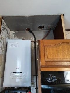 Chaudière murale blanche dans un meuble de cuisine, tuyau de gaz noir, meuble marron et four noir.