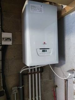 Une chaudière murale blanche avec un tuyau d'évacuation argenté, installée près de meubles en bois et d'une armoire.