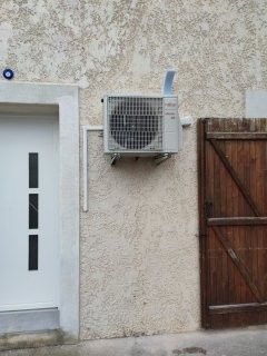 Unité de climatisation fixée sur un mur texturé de couleur claire, à côté d'une porte blanche et d'un portail en bois brun.