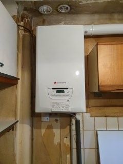 Chaudière murale blanche dans une cuisine, avec affichage numérique et tuyaux de raccordement.