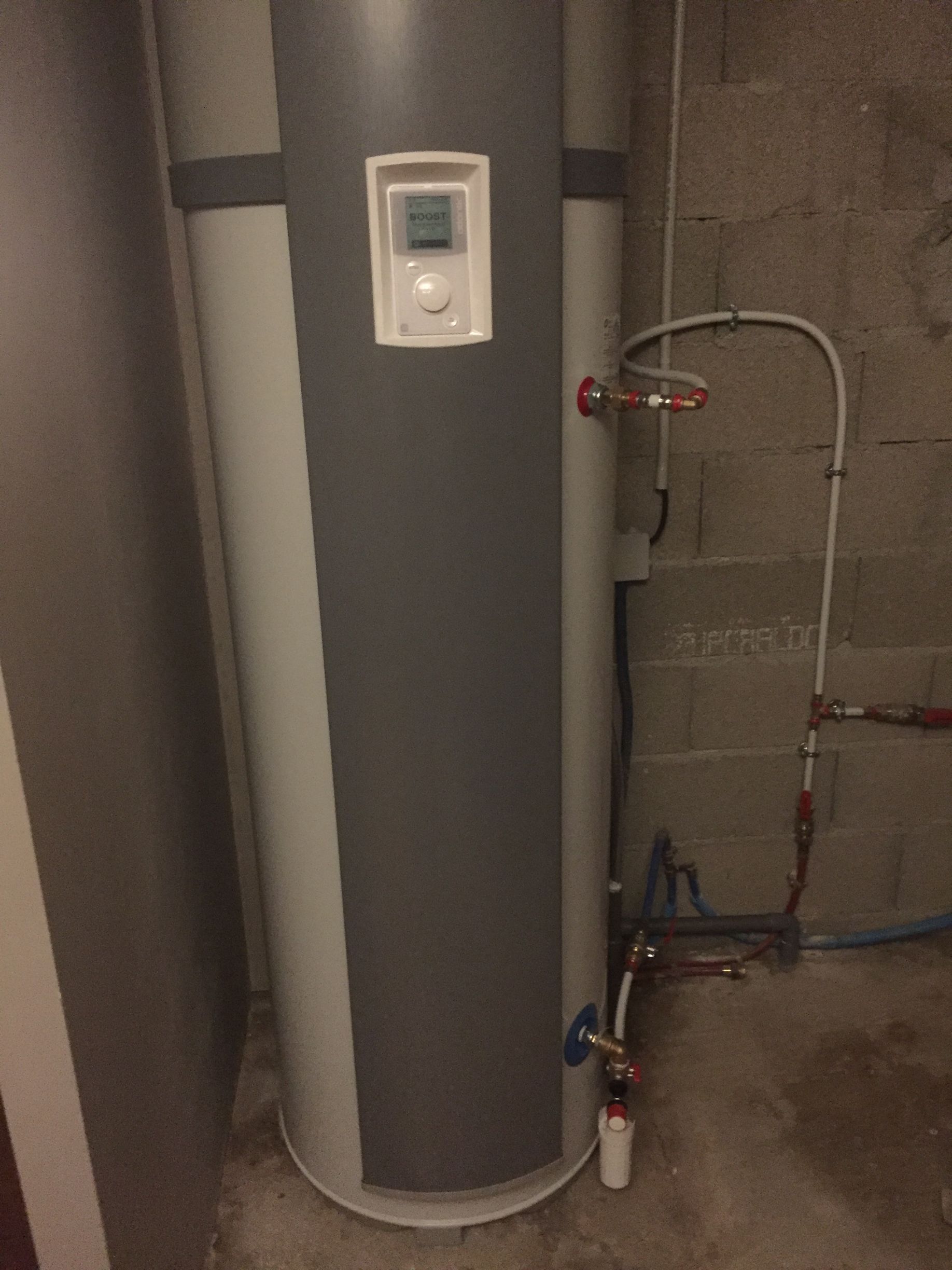 Un chauffe-eau cylindrique de grande taille, installé dans un sous-sol. Il est gris et blanc, avec des tuyaux raccordés et un affichage numérique.