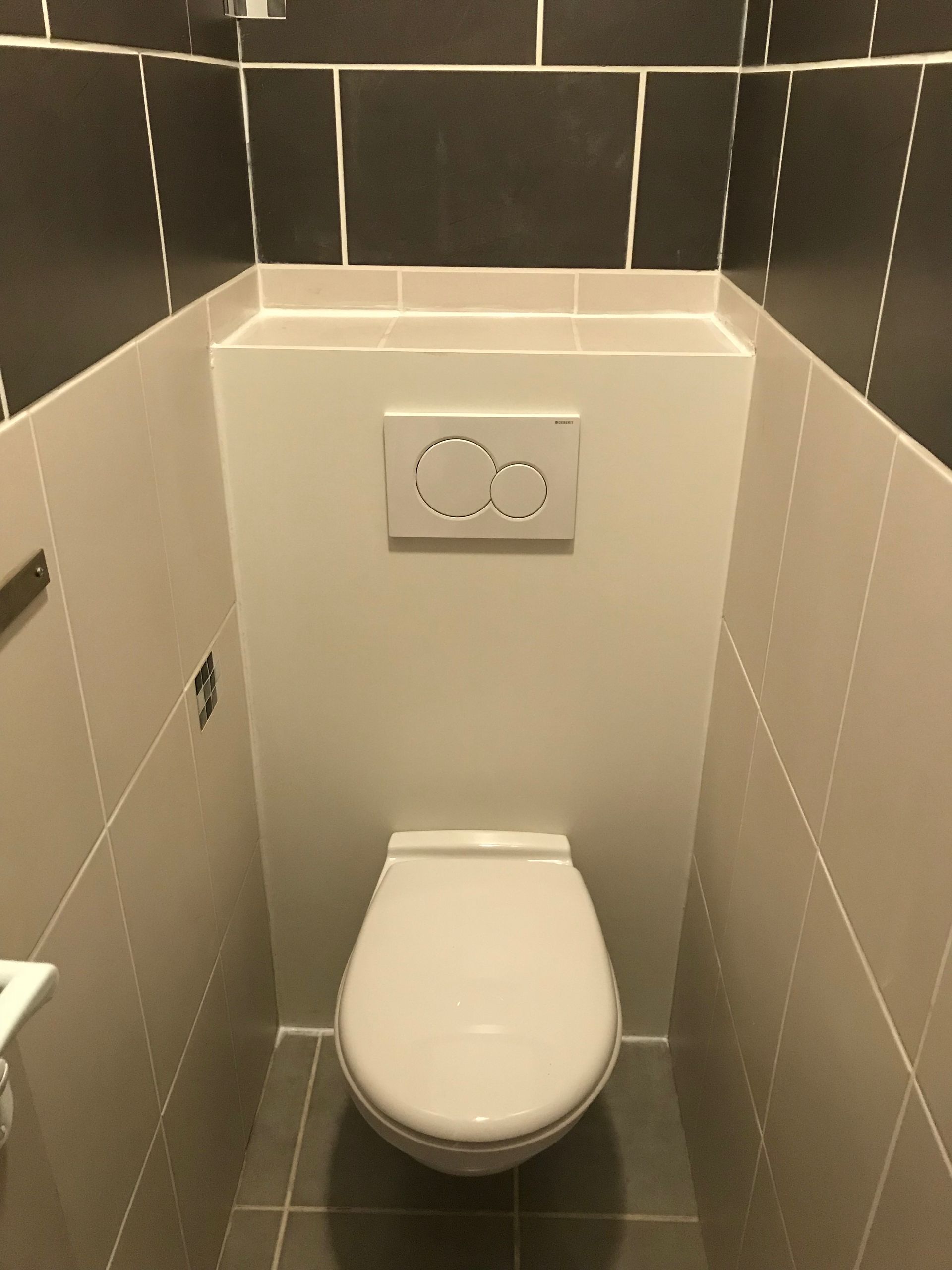 Toilettes dans une petite salle de bains carrelée. Abattant et bouton de chasse d'eau blancs. Carrelage gris et noir.