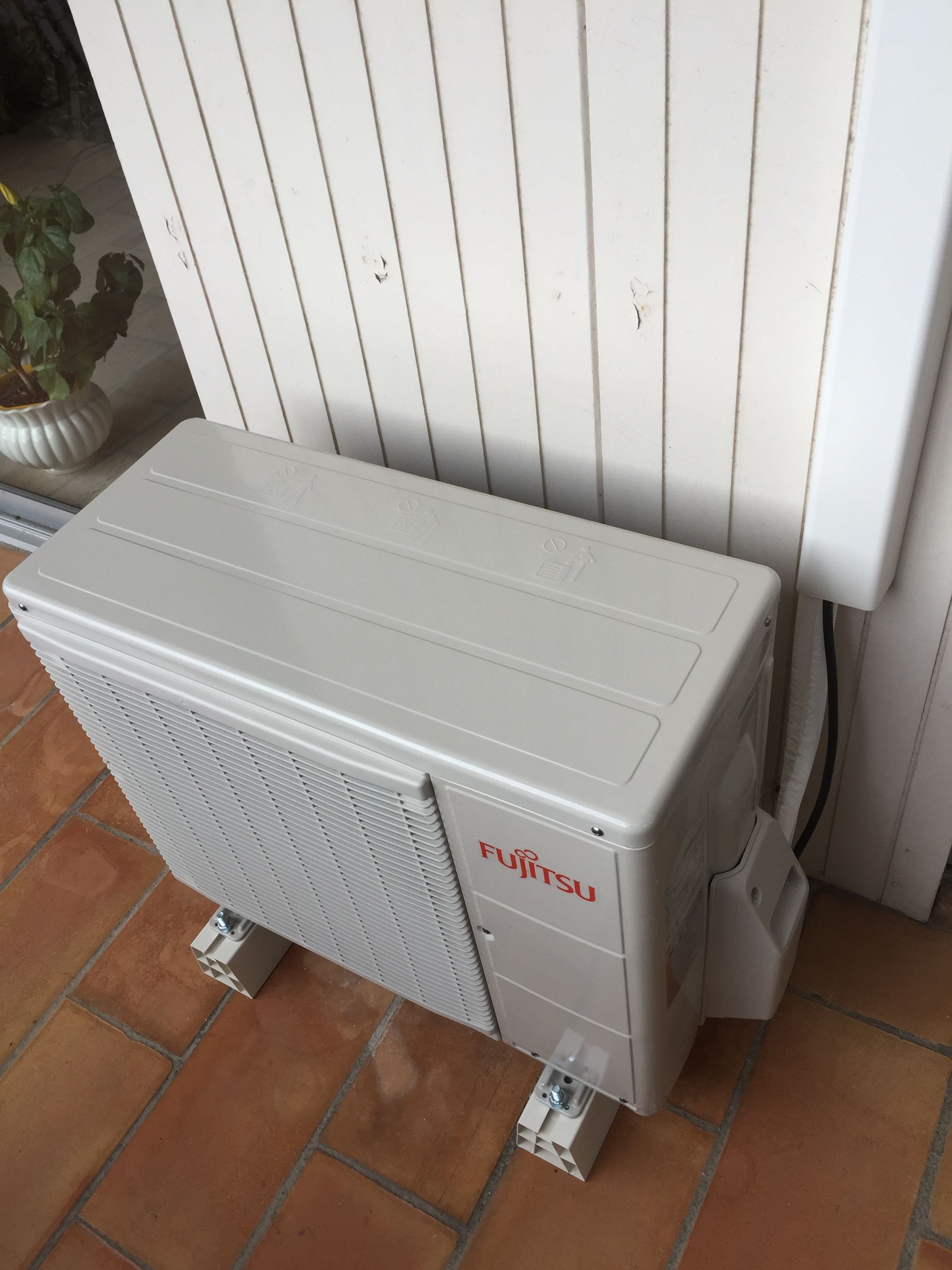 Unité de climatisation Fujitsu à l'extérieur, sur une terrasse en briques, contre un mur blanc.
