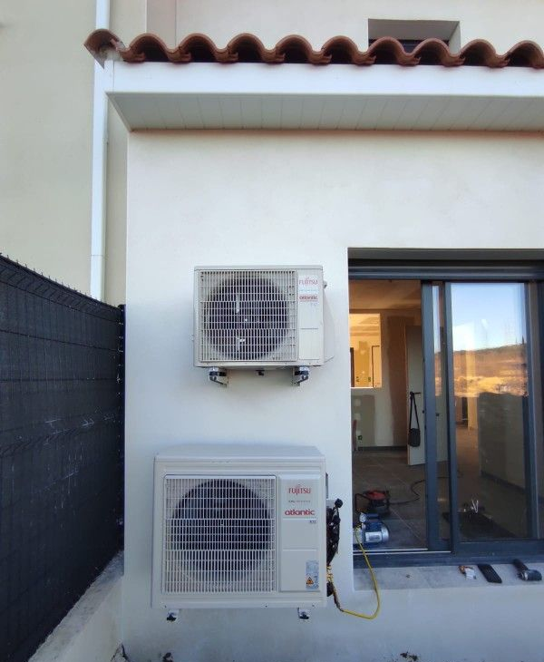 Deux unités de climatisation blanches sont fixées sur le mur extérieur d'un bâtiment. Une fenêtre se trouve à droite.