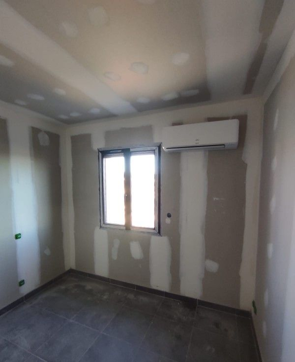 Pièce inachevée avec murs et plafond en plaques de plâtre, fenêtre, climatiseur. Le sol est foncé.