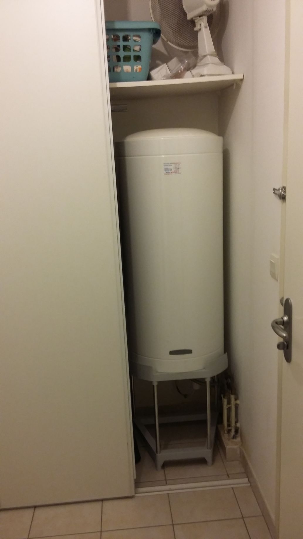 Un chauffe-eau blanc se trouve dans un petit espace clos, à côté d'un meuble. Un ventilateur et un panier à linge sont posés sur une étagère au-dessus.