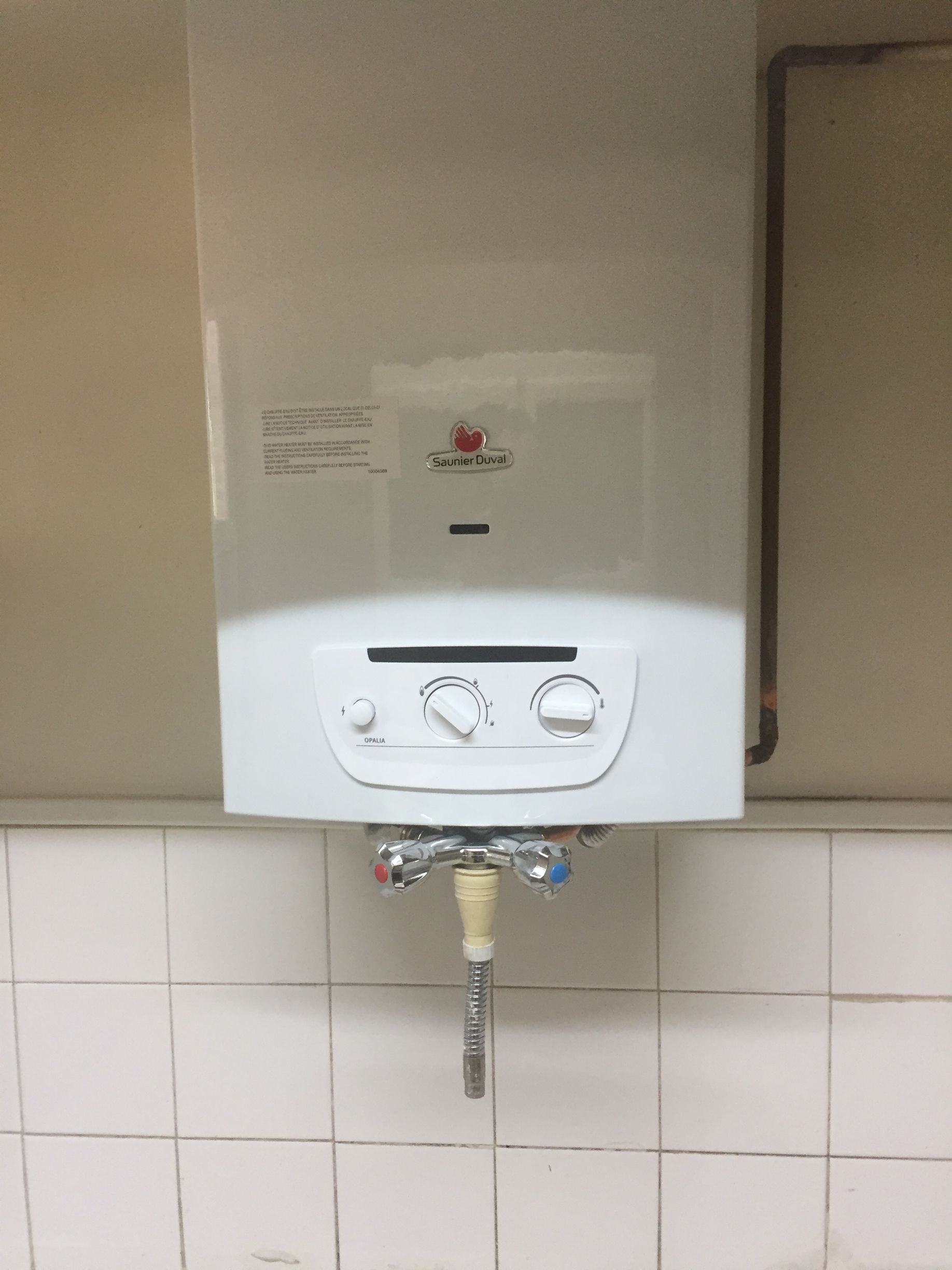 Chauffe-eau à gaz blanc fixé sur un mur carrelé. Commandes en dessous, tuyaux à droite.