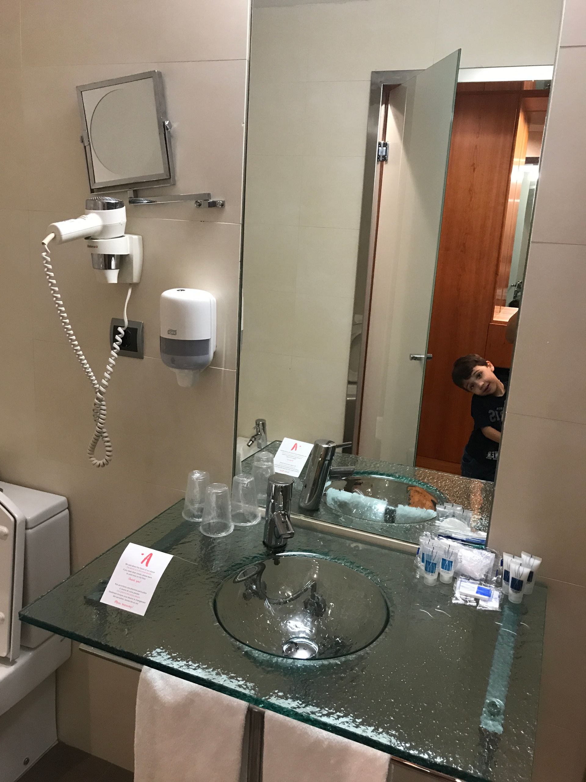 Salle de bains avec lavabo sur comptoir en verre, miroir et une personne qui regarde par l'embrasure d'une porte.