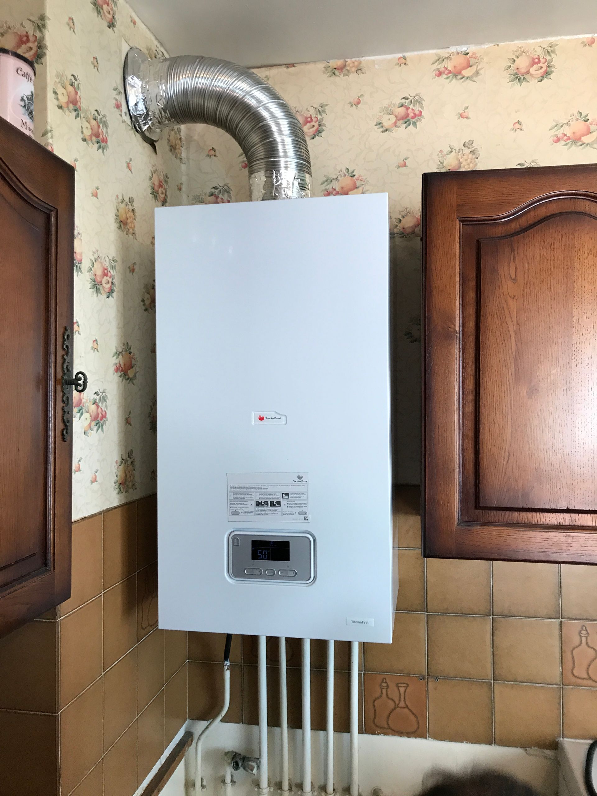 Chaudière murale blanche avec tuyau d'évacuation argenté, installée dans une cuisine aux murs carrelés et tapissés de papier peint à motifs floraux.