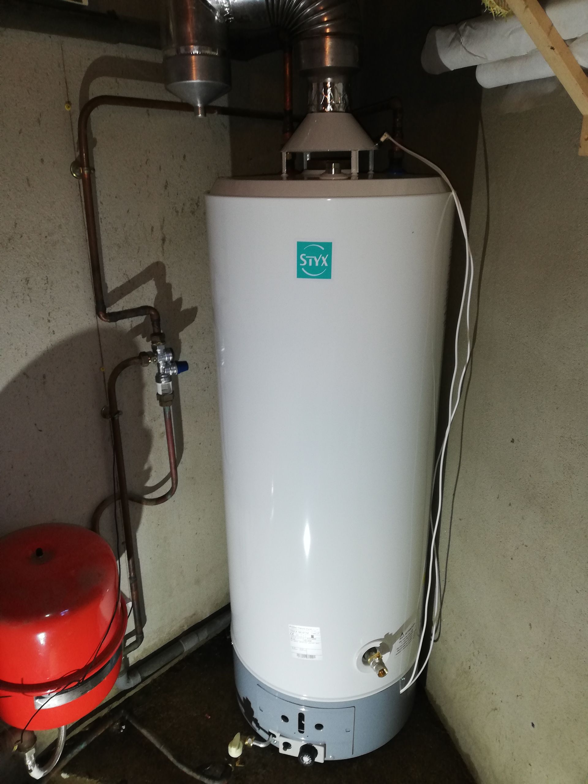 Chauffe-eau blanc dans un coin du sous-sol, raccordé à des tuyaux, à un conduit d'évacuation et à un vase d'expansion rouge.