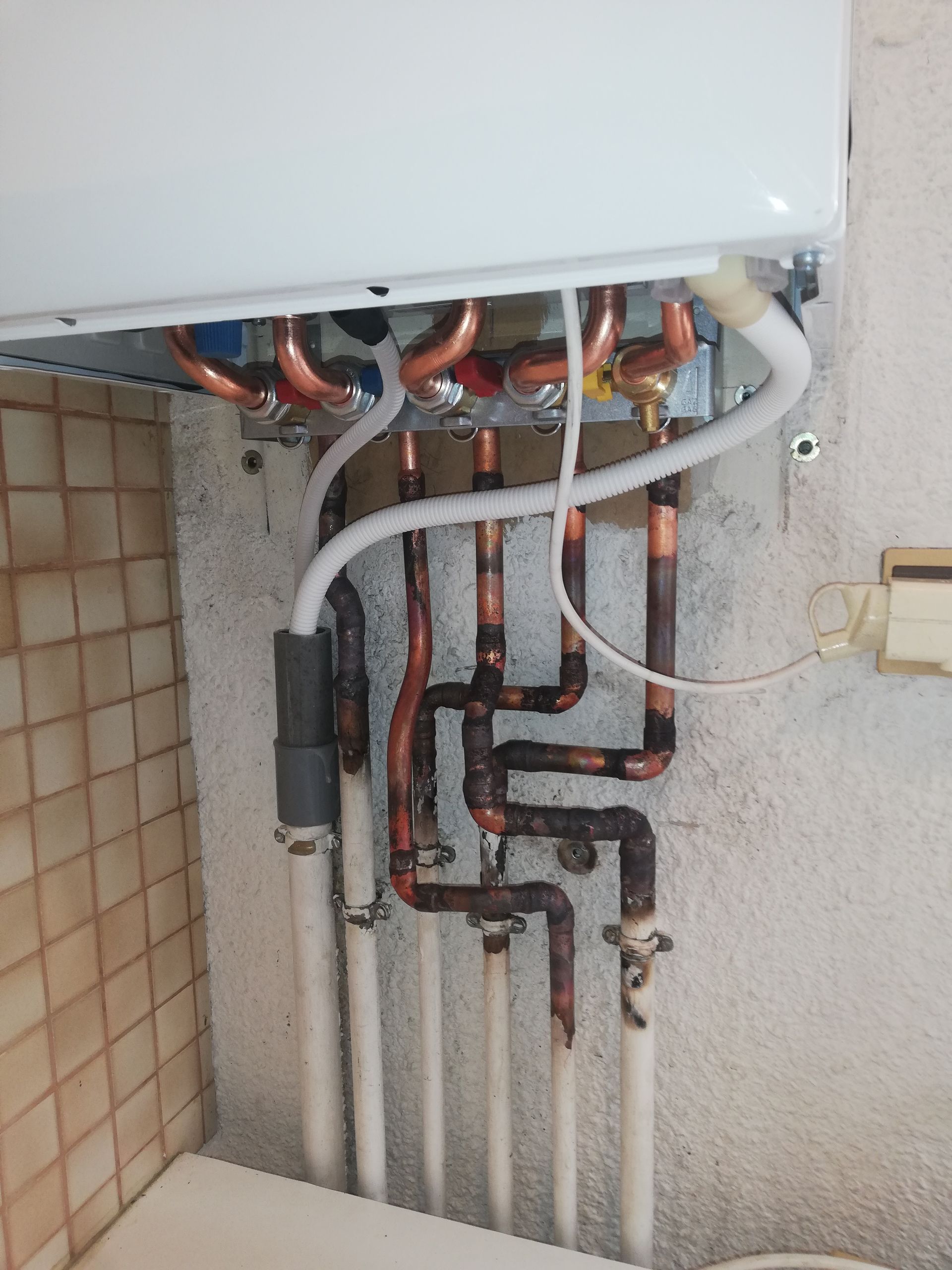 Des tuyaux en cuivre et des tubes en plastique blanc sont reliés à une chaudière blanche, fixée sur un mur texturé.
