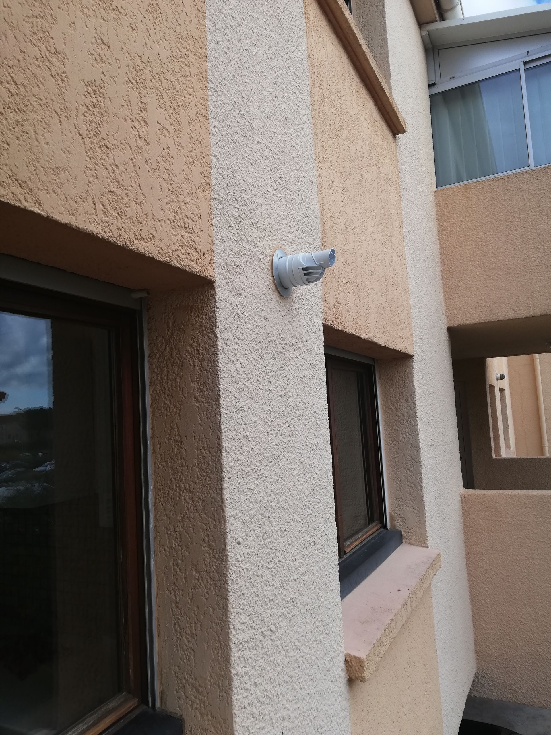 Extérieur du bâtiment avec caméra de sécurité fixée sur un mur beige texturé. Fenêtres à cadre foncé visibles.