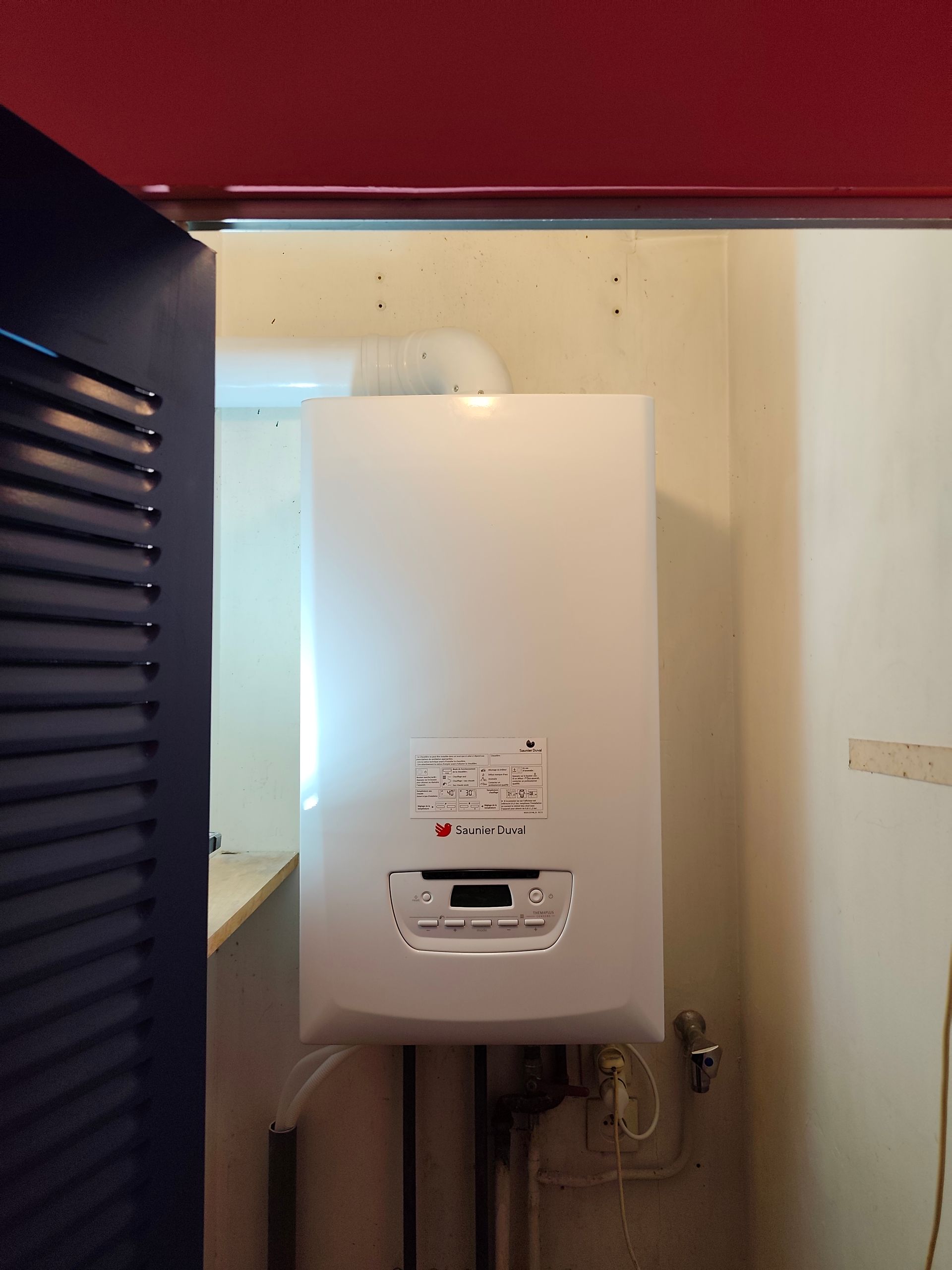 Chaudière murale blanche dans une buanderie, avec conduit d'évacuation, tuyaux et panneau de commande visibles.