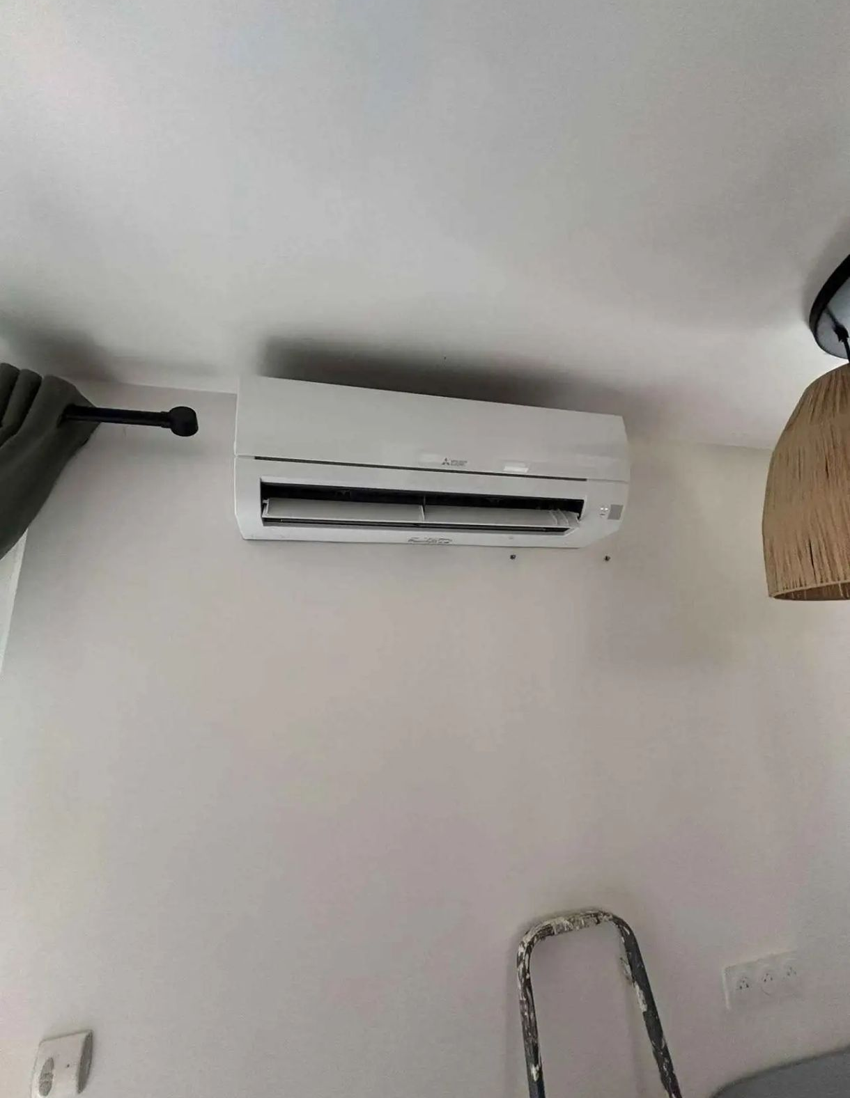 Un climatiseur blanc sur un mur blanc ; une tringle à rideaux foncée et une suspension lumineuse sont partiellement visibles.