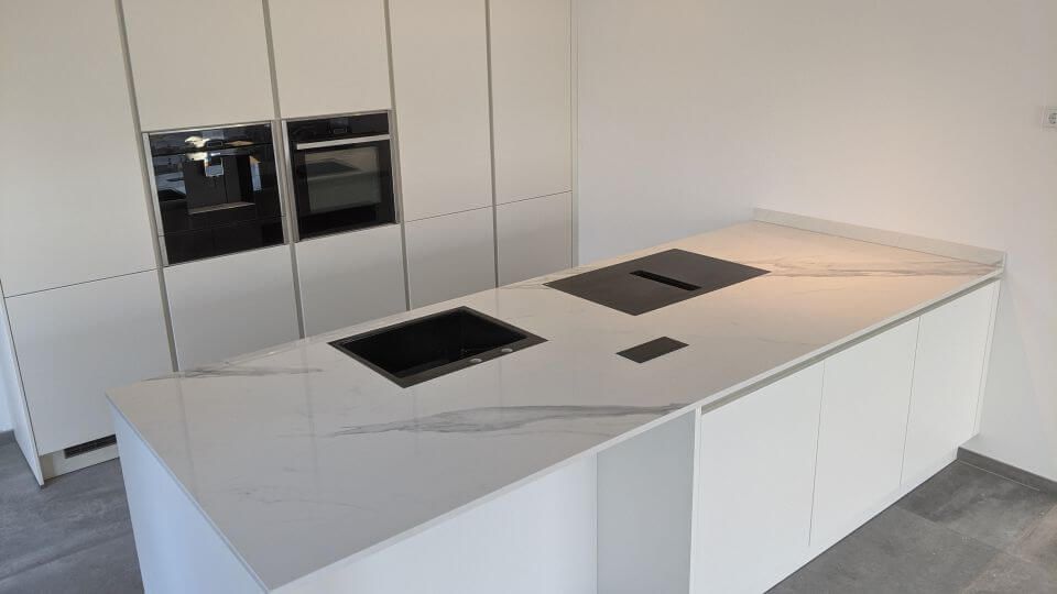 Moderne weiße Kücheninsel mit eingebautem Kochfeld und dunklen Arbeitsflächenöffnungen, daneben hohe weiße Schränke.