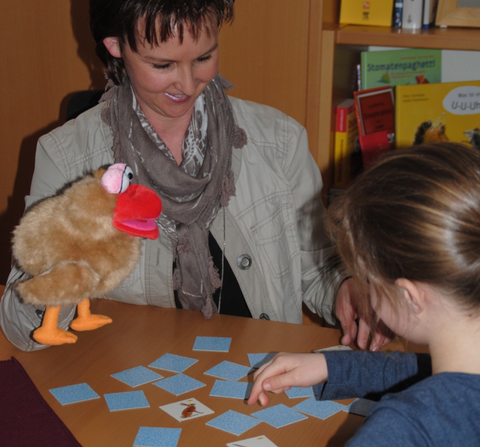 Frau spielt mit einer Puppe und einem Kind an einem Tisch ein Memory-Spiel.