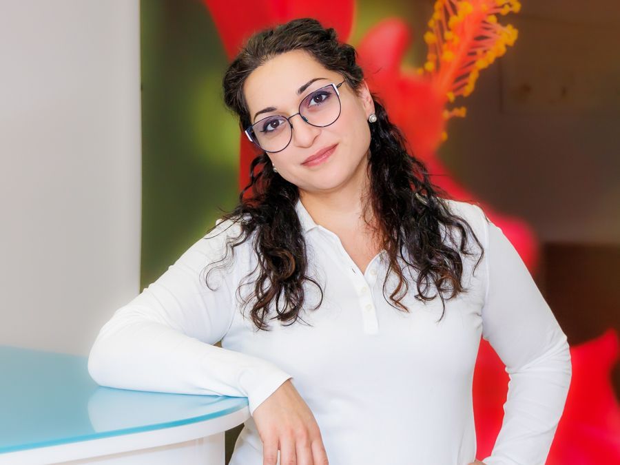 Eine Person mit lockigem, dunklem Haar und Brille trägt ein weißes, langärmeliges Hemd und lehnt an einem Schreibtisch.