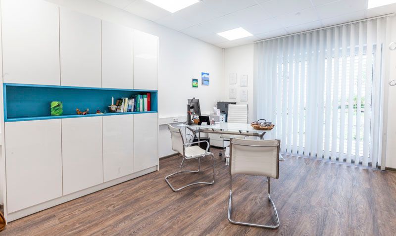 Ein modernes Büro mit weißen Schränken, einem türkisfarbenen Regal, zwei weißen Stühlen und einem Schreibtisch.