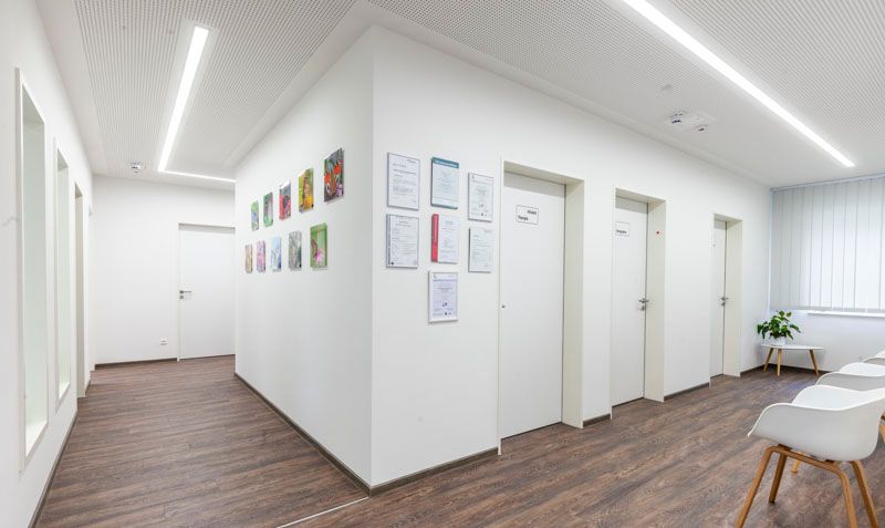 Ein moderner Wartebereich in einer Arztpraxis mit weißen Wänden, Holzbodenoptik, gerahmten Urkunden und weißen Stühlen.