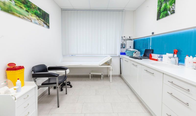 Ein sauberer, moderner Untersuchungsraum mit weißer Untersuchungsliege, Patientenstuhl und weißen Schränken.