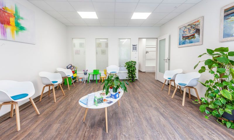 Ein Wartezimmer mit weißen Stühlen, einem ovalen Tisch in der Mitte, Topfpflanzen und Wandbildern auf hellem Holzboden.