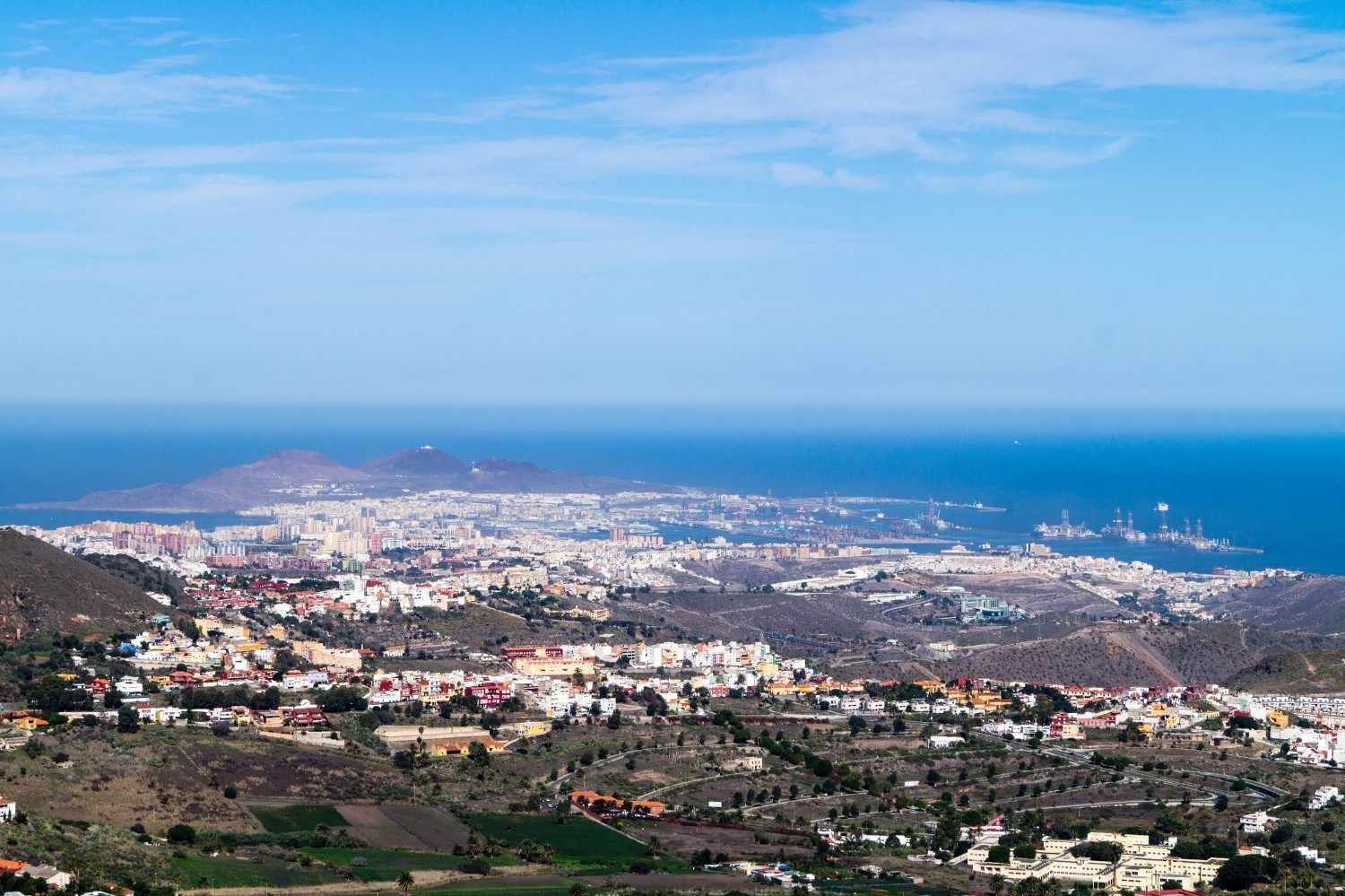 Ciudad costera y puerto vistos desde una ladera bajo un cielo azul despejado.