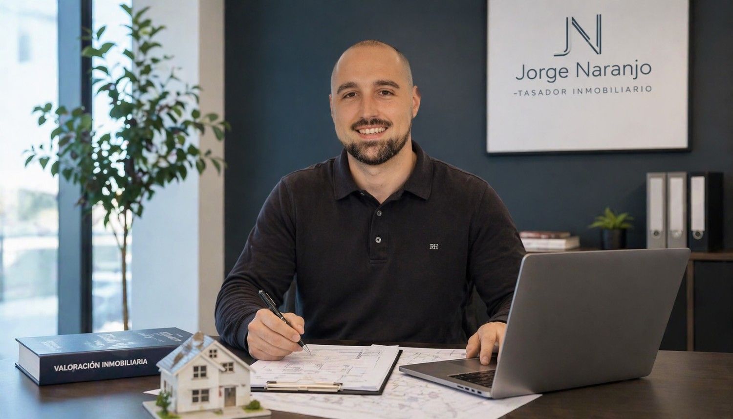 Un hombre sentado en un escritorio con una computadora portátil y un cuaderno en una oficina, sonriendo junto a un letrero que dice 