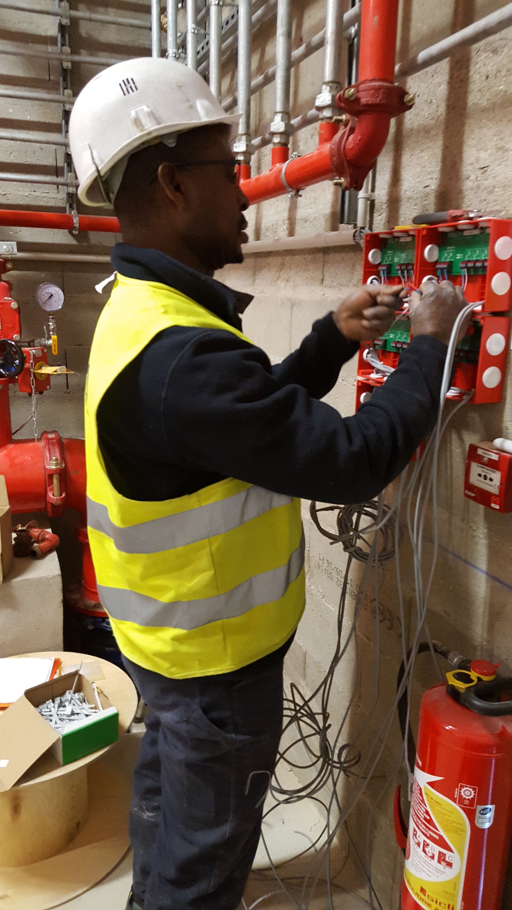Un technicien, portant un casque et un gilet de sécurité, travaille sur le câblage électrique d'un panneau de commande dans un local technique industriel.
