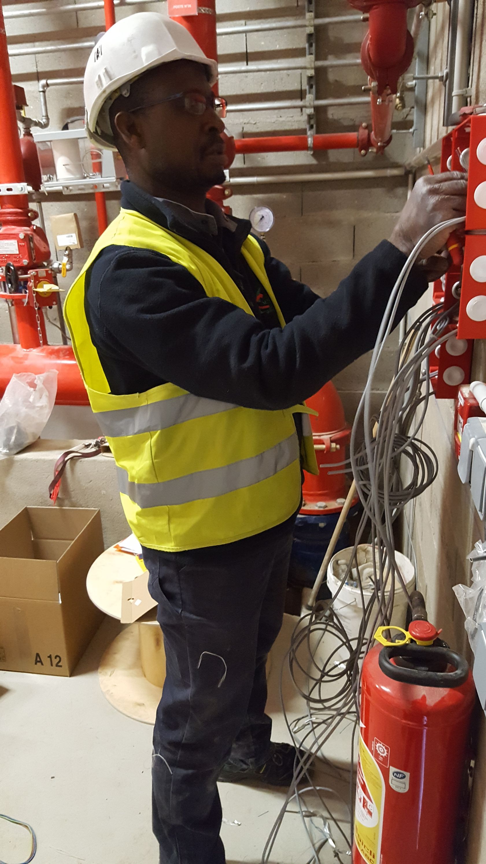 Un ouvrier portant un gilet haute visibilité et un casque de chantier effectue le câblage d'un tableau électrique dans un local technique industriel.