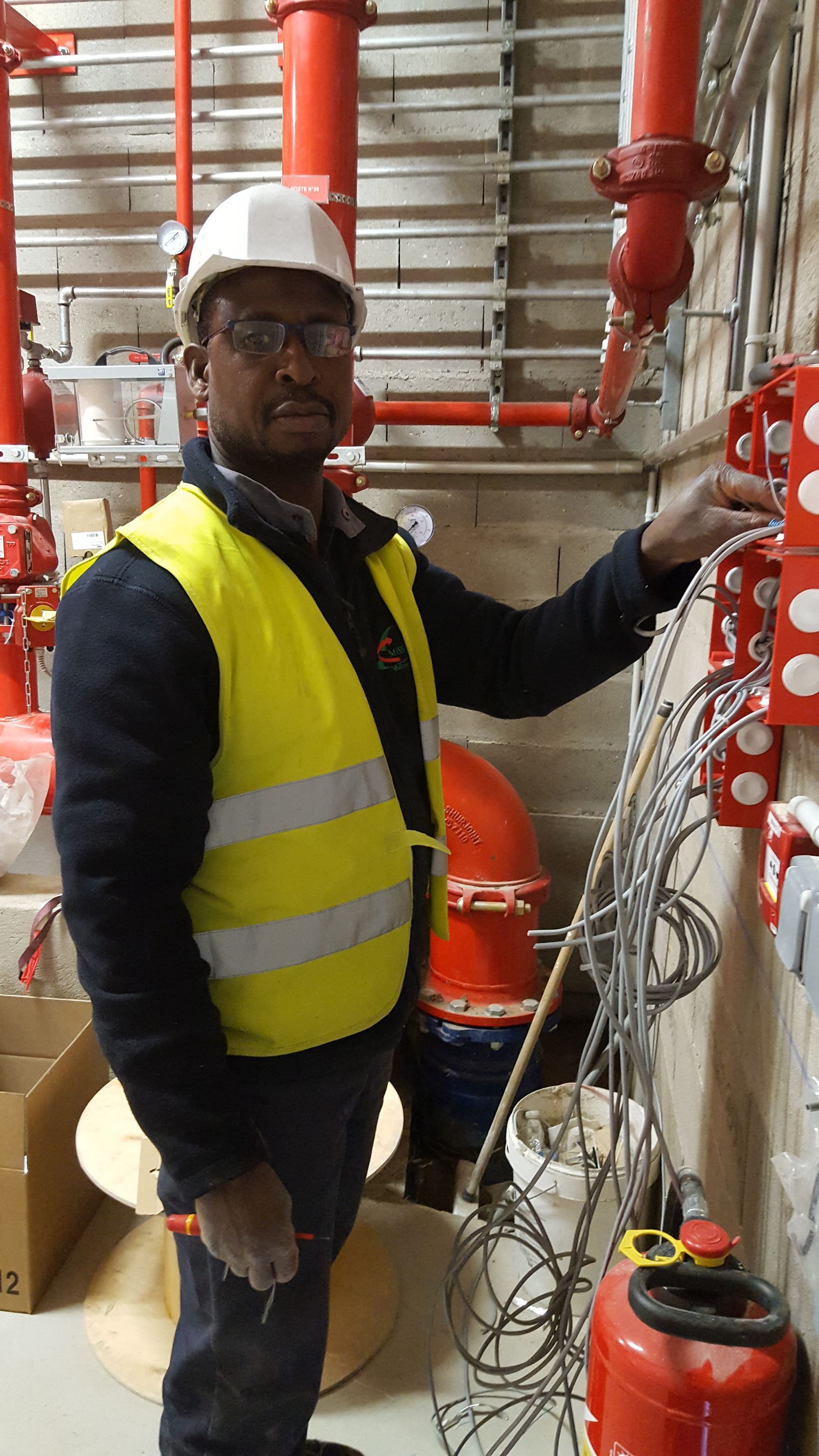 Un ouvrier portant un gilet haute visibilité et un casque de chantier inspecte le câblage électrique sur un mur d'un local technique, avec des tuyaux.