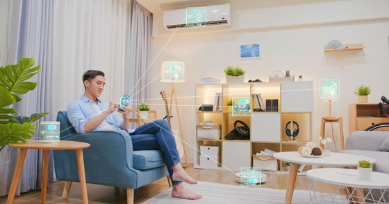 Une personne est assise dans un fauteuil, dans un salon, et utilise un smartphone pour gérer des appareils domotiques connectés.