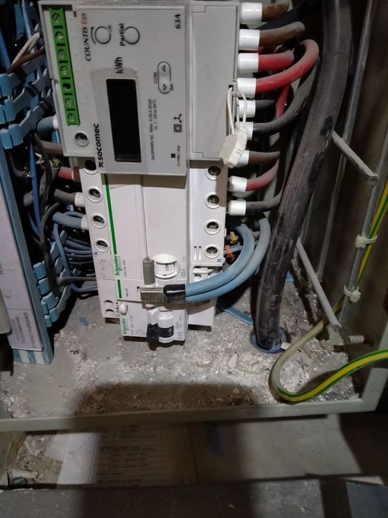 Un tableau de distribution électrique avec un compteur d'énergie numérique, des câbles connectés et un fil de terre, le tout dans un boîtier poussiéreux.