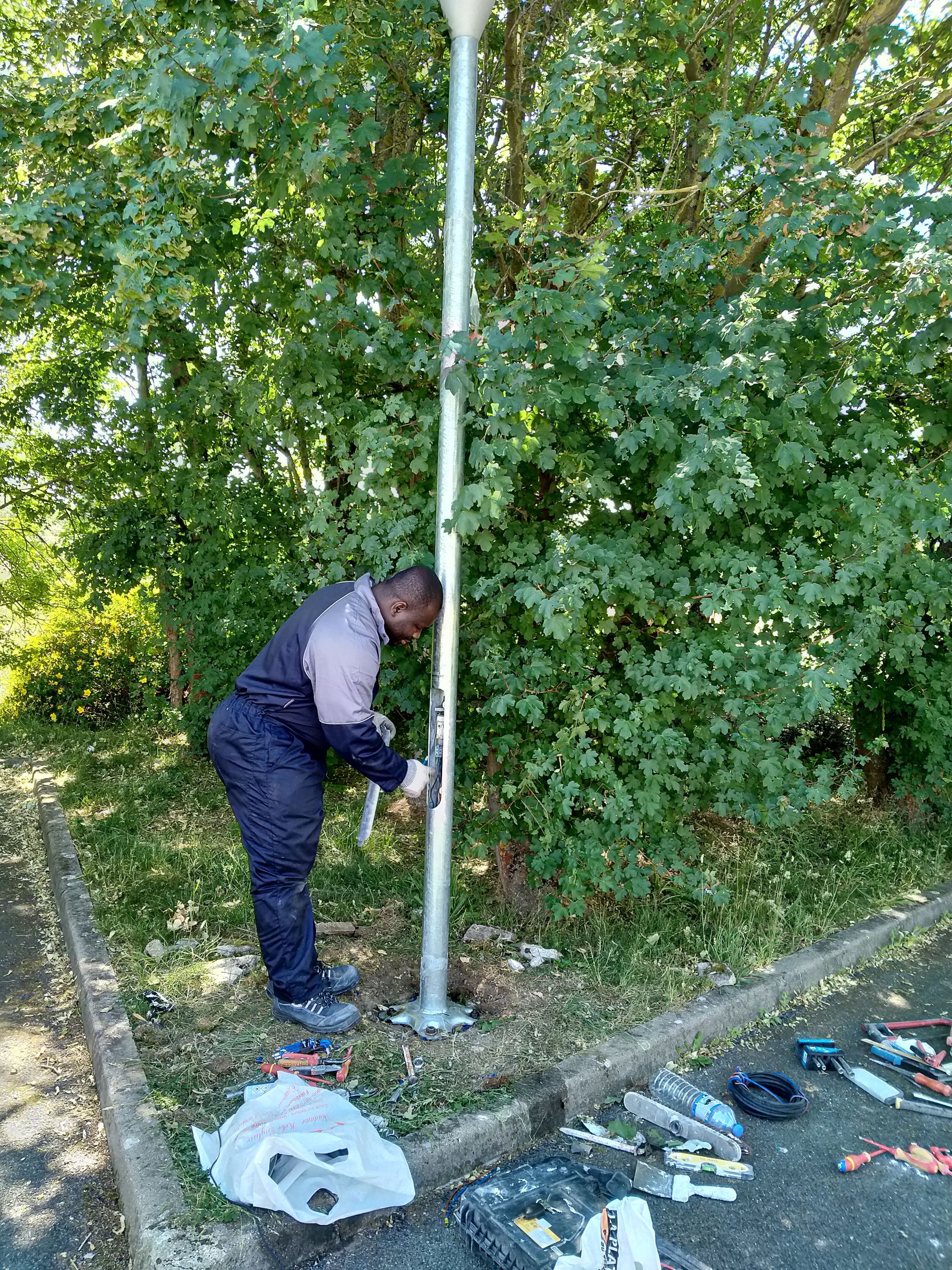 Une personne en uniforme peint un poteau métallique qui se dresse au bord d'une route, près d'arbres verdoyants. Des outils sont posés sur le trottoir.
