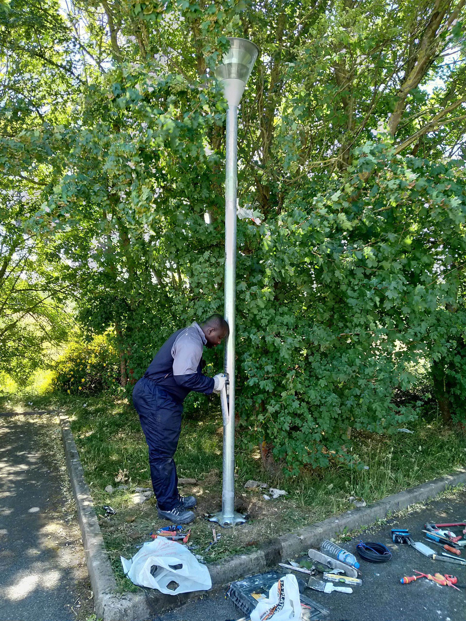 Une personne en uniforme fixe un poteau métallique à l'extérieur, près d'un trottoir bordé d'arbres et jonché de détritus.