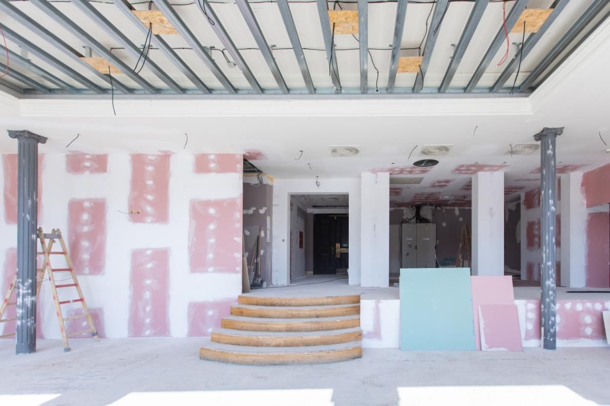 Espacio interior en construcción; estructura de techo expuesta, paneles de yeso rosa, escalones, columnas grises.