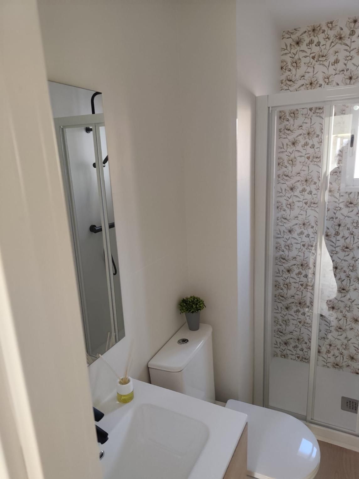 Baño con inodoro blanco, lavabo, ducha con papel pintado floral.