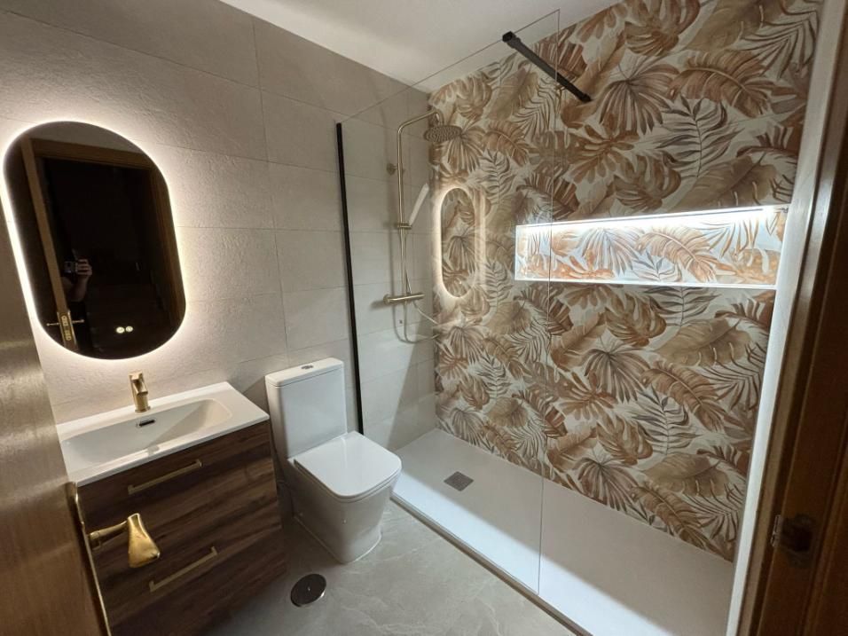 Baño moderno con espejo ovalado, tocador de madera, inodoro y ducha con pared estampada.