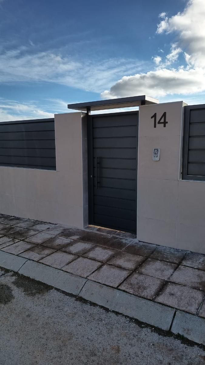 Puerta gris en una pared de color rosa claro, con el número de dirección 14. Cielo nublado al fondo.