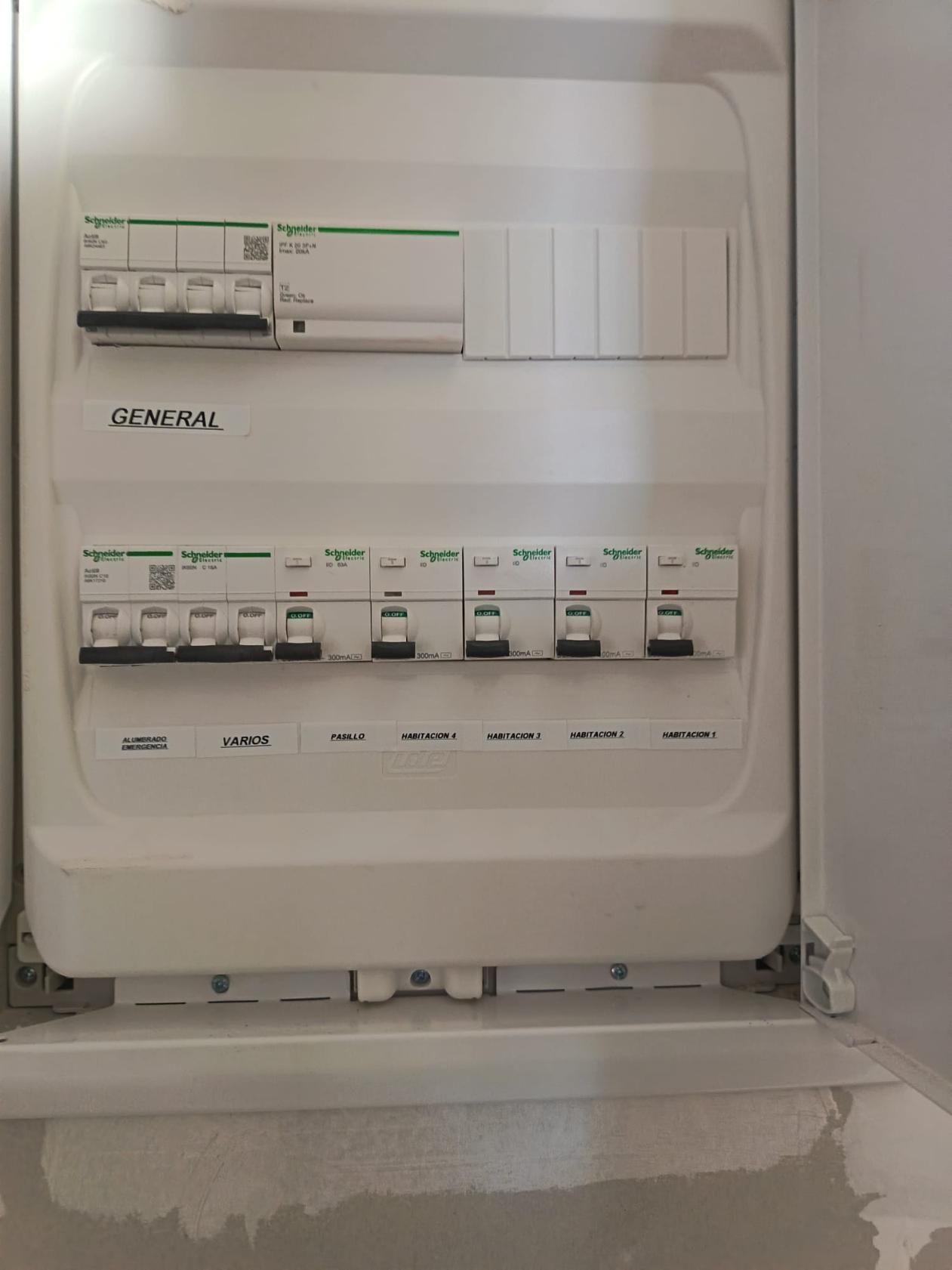 Panel eléctrico blanco con múltiples disyuntores e interruptores etiquetados, montado en una pared.
