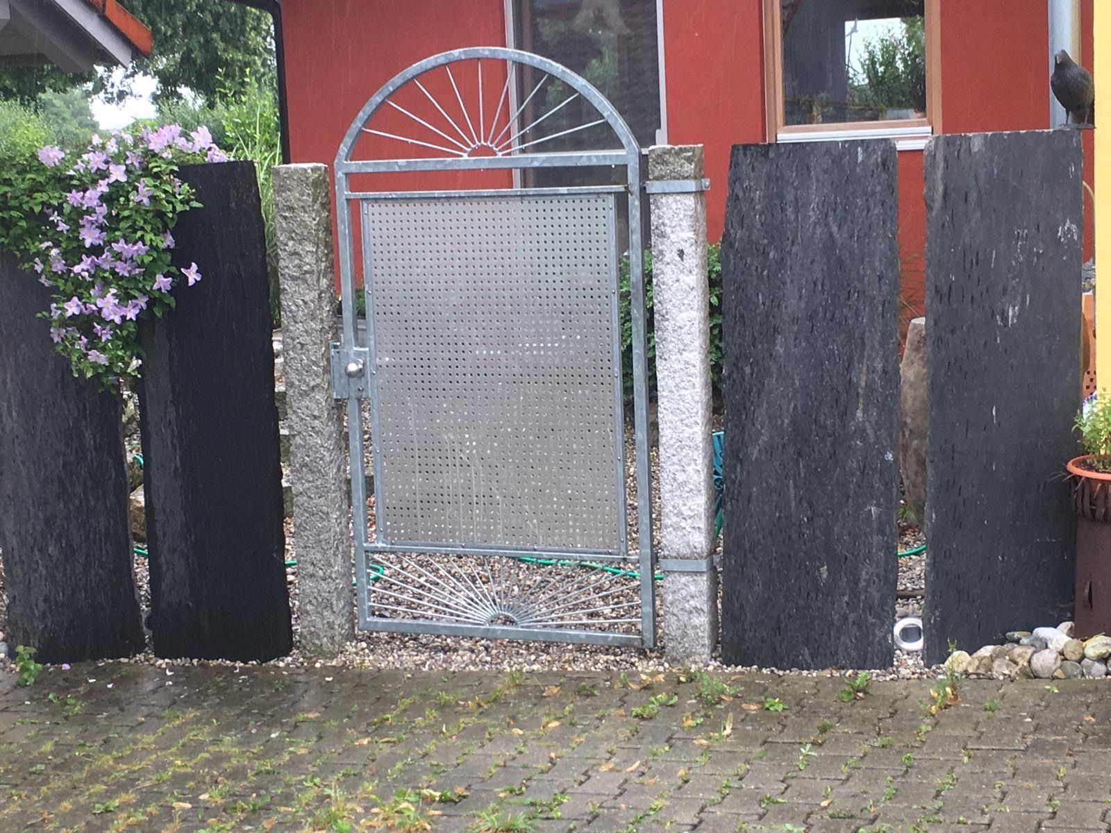 Ein Metalltor zwischen schwarzen Kiespfosten mit Blumen, vor dem Eingang eines roten Hauses