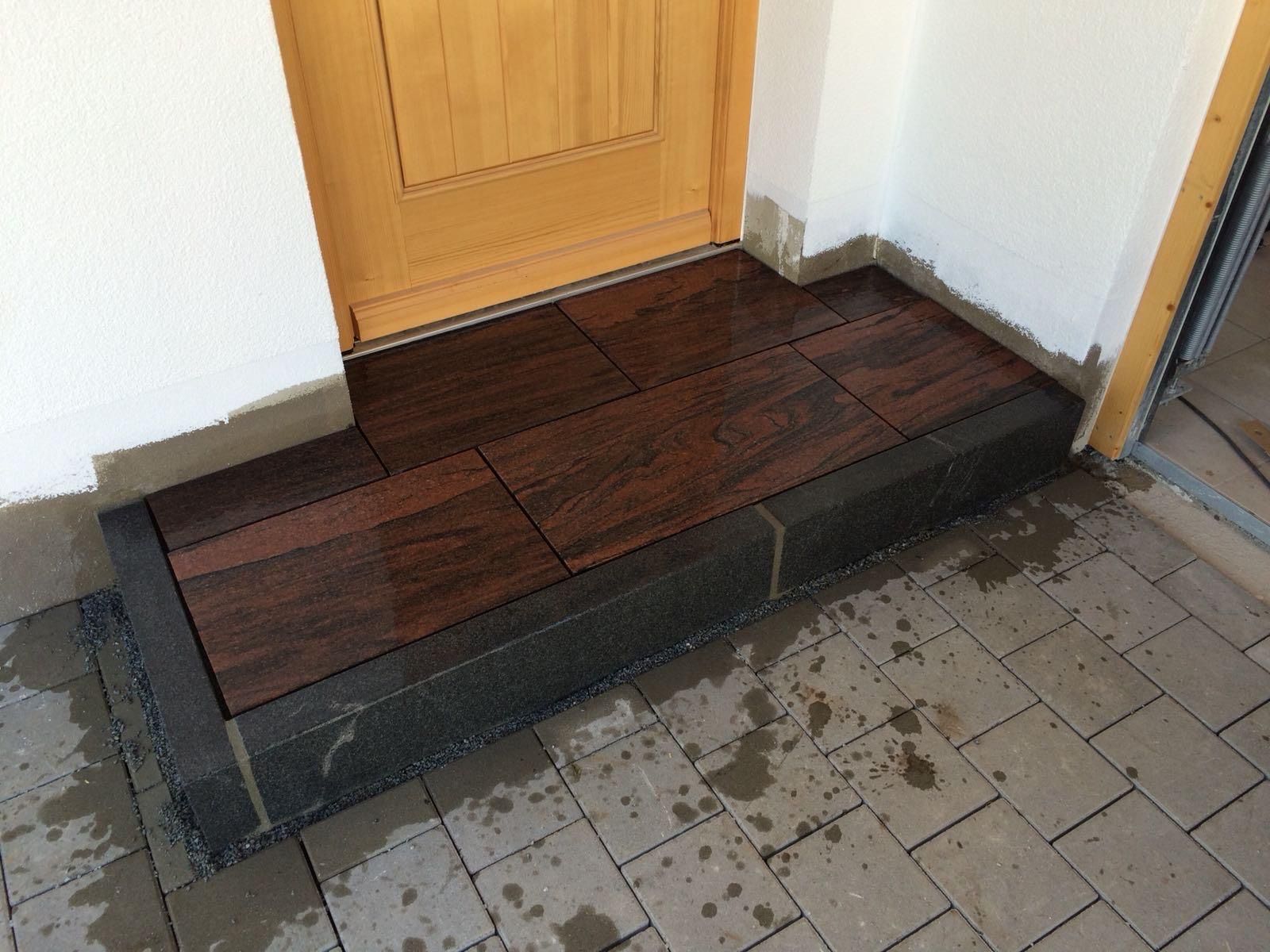 Erhöhte Türschwelle mit dunkler Holzrampe, die zu einer gelben Tür und einem gefliesten Gehweg führt.