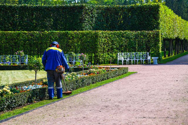 Eine Person in blauer Jacke und blauem Hut steht neben einem Blumenbeet in einem gepflegten Gartenlabyrinth.