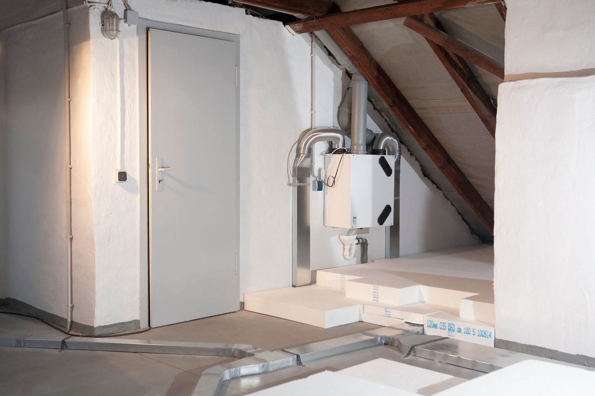 Intérieur d'un grenier avec une porte grise fermée, un système de ventilation et des poutres apparentes. Murs blancs.