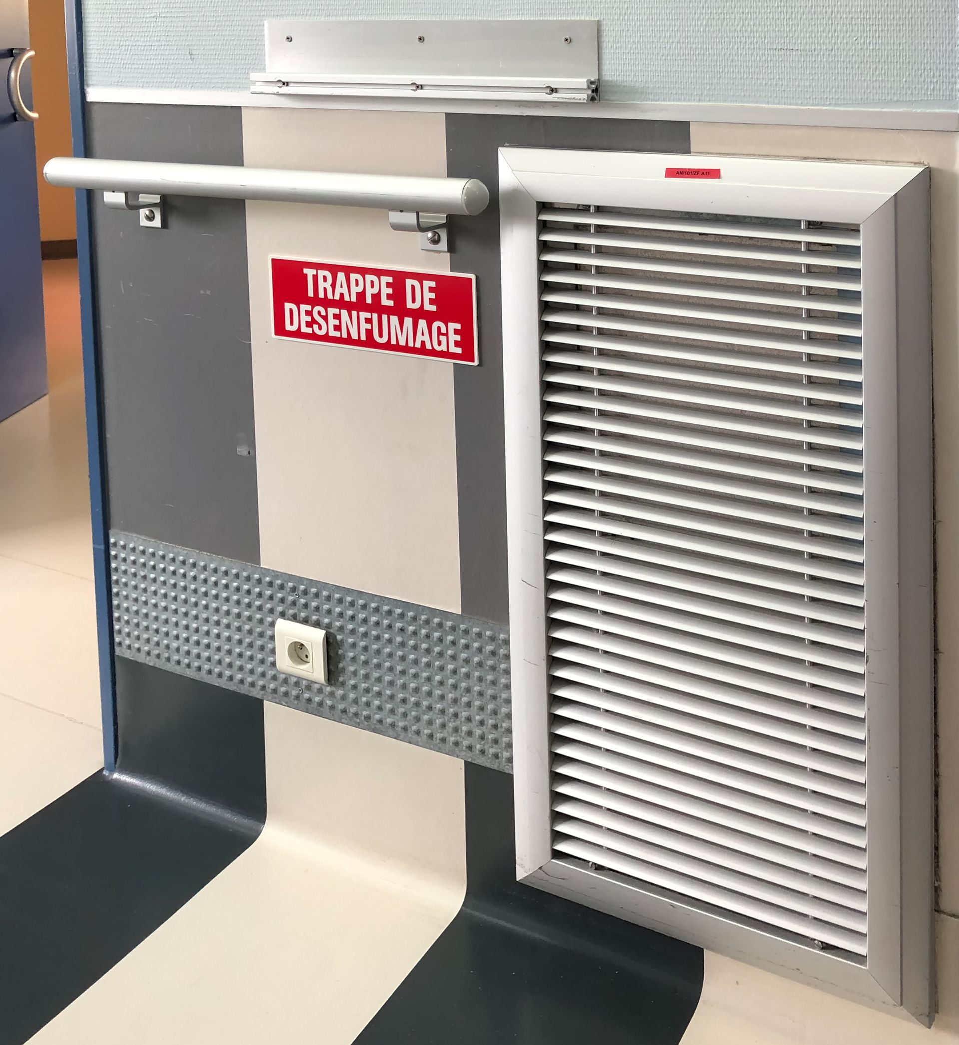 Mur avec conduit d'évacuation des fumées, rambarde et panneau en français. Couleurs : gris, blanc et bleu.