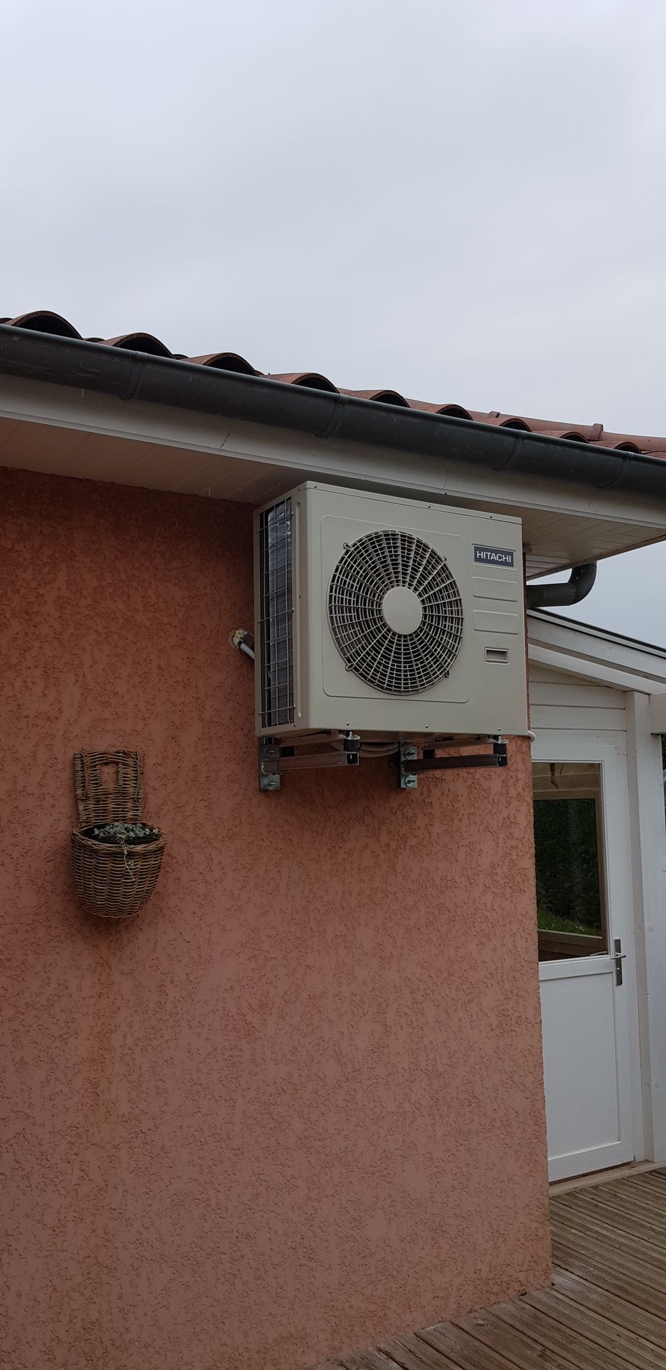 Unité de climatisation fixée sur un mur extérieur couleur pêche, à côté d'une porte blanche. Ciel couvert.