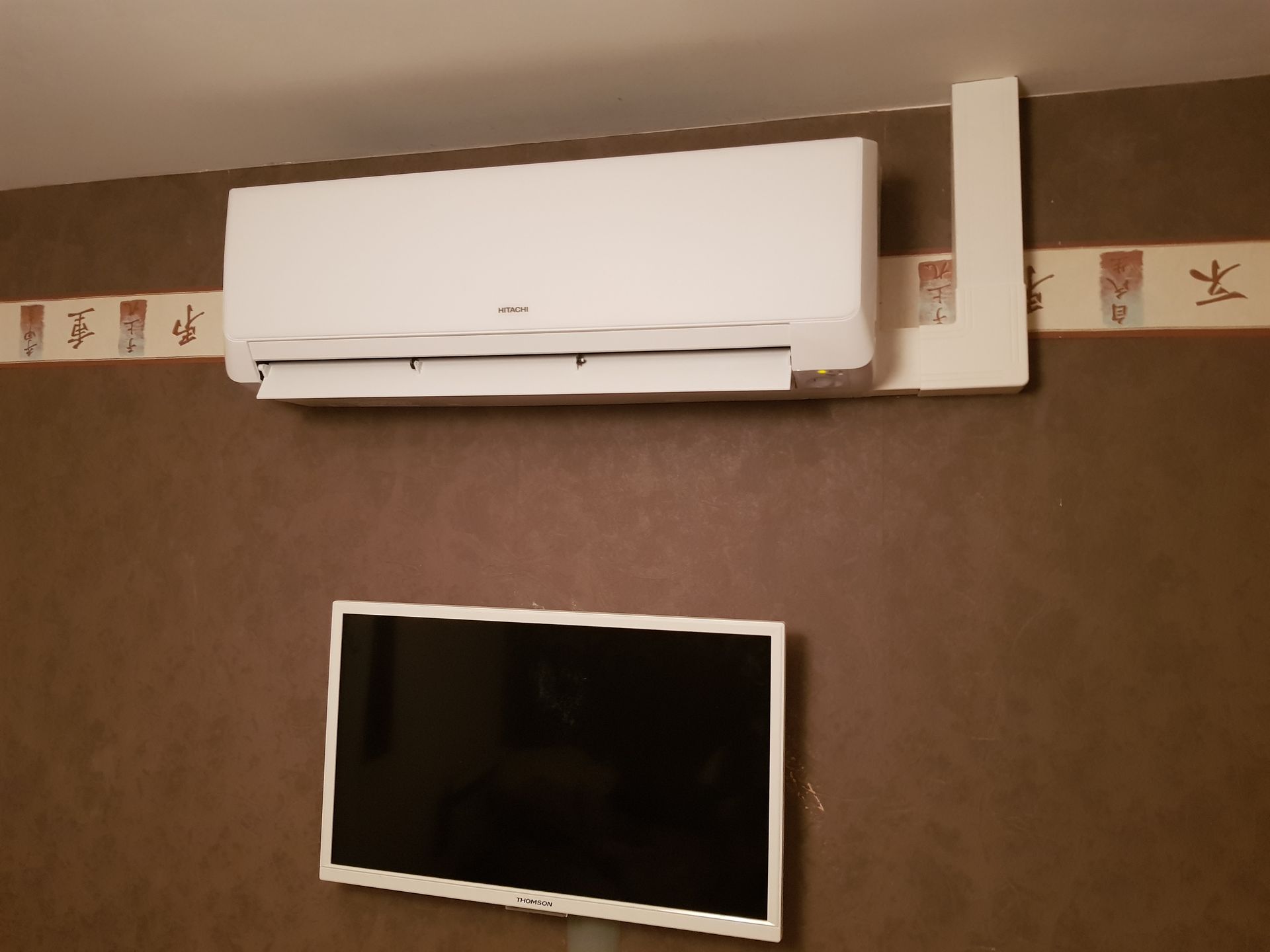 Un climatiseur blanc est placé au-dessus d'un téléviseur fixé au mur, sur un mur marron orné d'une bordure décorative.